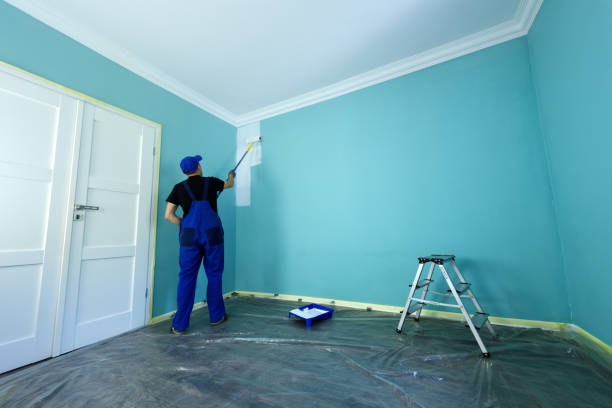 Man in a working overall is painting the wall in white color